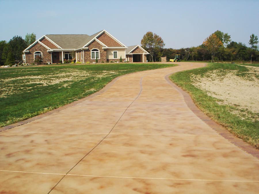 A curving stamped concrete driveway leads to a large single-story residential home with a prominent front yard. The house has a brick facade with white trim around the windows, and a well-manicured lawn. The driveway is bordered by undeveloped land, suggesting a rural or suburban setting with ample space and a peaceful environment.