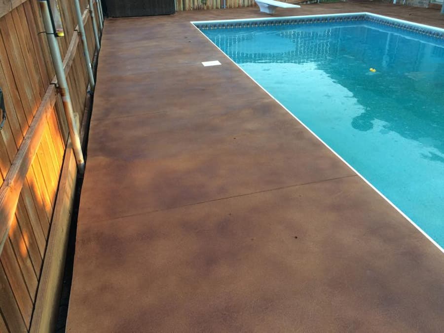 A poolside area with a smooth, stained concrete deck in a rich, rust-brown color, complementing the blue waters of the swimming pool. The edge of the pool is lined with white tiles, providing a neat border. A wooden fence runs alongside the deck, creating a private and serene backyard oasis. The surface of the water reflects a gentle ripple, indicating a tranquil setting.