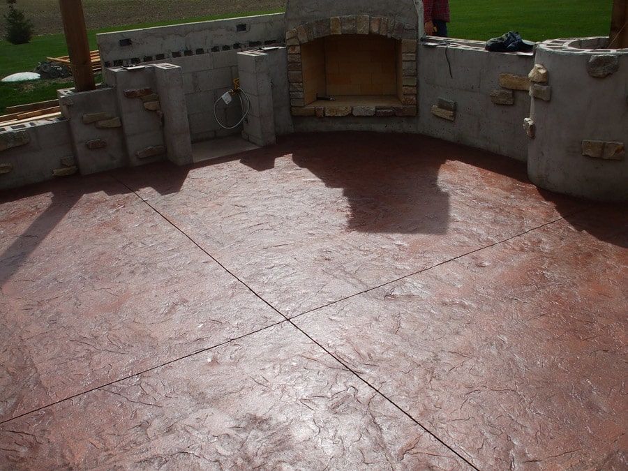 A patio area with a textured, rose-colored stamped concrete floor. The patio is part of an outdoor living space featuring a half-built stone fireplace and an in-progress outdoor kitchen with block construction. The open backyard visible in the background and the construction materials suggest ongoing work to create an inviting outdoor entertainment area.