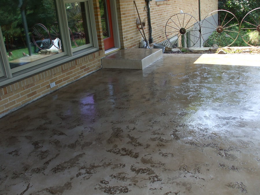 A newly finished concrete patio adjacent to a brick house with large windows reflecting the surrounding trees. The surface of the patio has a mottled pattern with darker patches, likely from a recent rain as evidenced by the wet sheen. In the background, decorative antique metal wheels are mounted on the wall, adding to the rustic charm of the outdoor space.