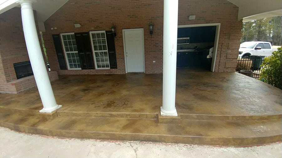 A spacious covered patio area with a stained concrete floor, blending shades of brown and tan. The patio features white columns supporting the roof, a brick exterior wall with French doors and a single white door. An outdoor fireplace is built into the brick wall, and a part of a white pickup truck is visible in the driveway, indicating a residential setting.