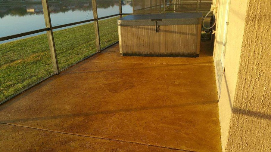 A golden-hued, textured concrete patio extends towards a screened enclosure overlooking a tranquil lakeside. The early morning or late afternoon sunlight bathes the area in a warm glow, highlighting the smooth surface of the patio. A hot tub sits at the edge, promising relaxation with a view of the serene water and lush green lawn beyond the screen.