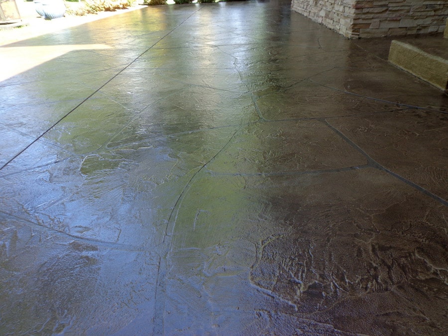 The photo showcases a spacious stamped concrete patio with a glossy finish that reflects the surroundings. The patio is designed with varying shades of green and brown, creating a stone-like appearance. The wet surface indicates it has either been cleaned or rained on recently. Part of a stone structure is visible on the right, adding to the patio's rustic charm.
