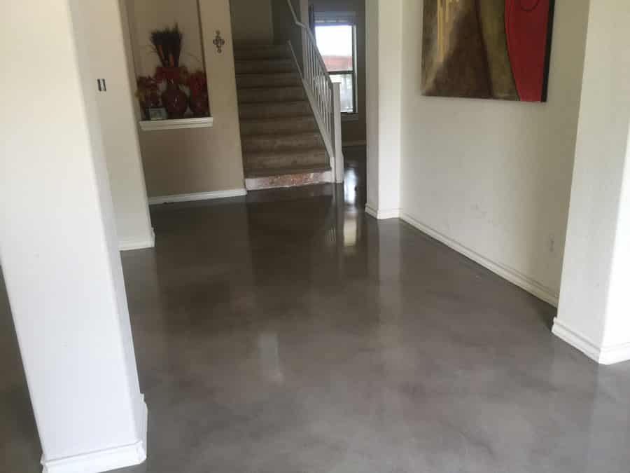 An entryway in a modern home featuring a polished concrete floor with a subtle marbled effect. The space is bright and airy, with white walls complementing the floor's neutral tones. To the right, a staircase with a carpeted runner leads to the upper level, and decorative elements like a vase and wall art add color and personal touches to the welcoming area.