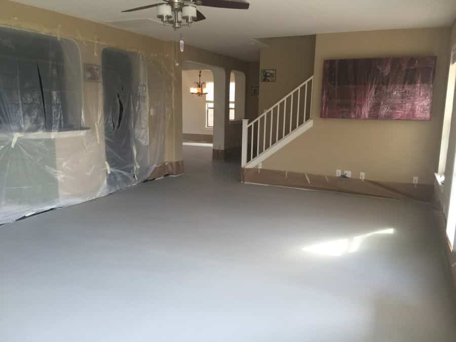 An interior room during renovation with a freshly coated gray epoxy floor that provides a smooth, reflective surface. The room is well-lit, with natural light streaming in, casting soft shadows. The walls are protected with plastic sheeting, and a white staircase with a wooden handrail leads to another level. A large, abstract painting hangs on one wall, adding a pop of color to the otherwise neutral palette of the space.